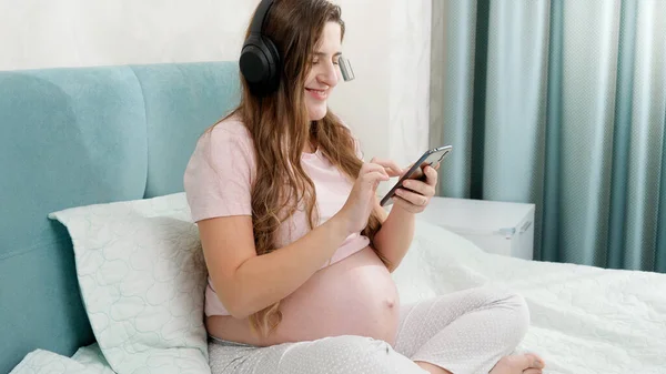 Feliz sorrindo grávida ouvindo música com fones de ouvido e usando smartphone enquanto está sentado na cama em casa — Fotografia de Stock