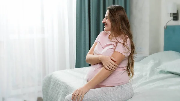 Beautiful pregnant woman in pajamas sitting on bed at morning and looking on sun shining through window — Stock Photo, Image