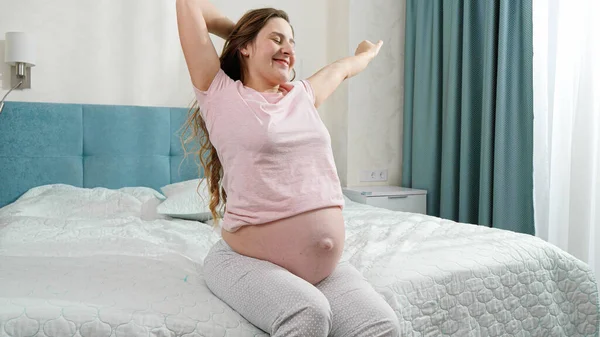 Happy smiling pregnant woman in pajamas waking up in morning and looking out of the window. Concept of happiness during pregnancy and expecting baby — Stock Photo, Image