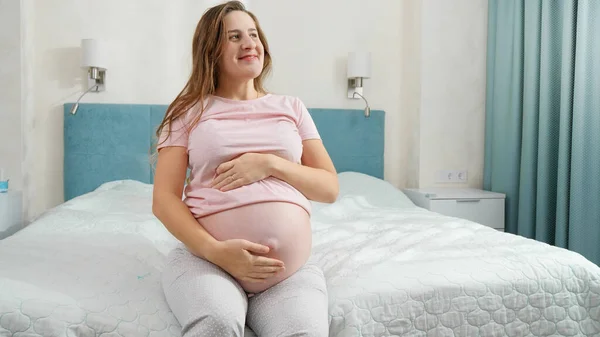 Porträtt av lycklig leende gravid kvinna i pyjamas sitter på sängen på solig morgon — Stockfoto