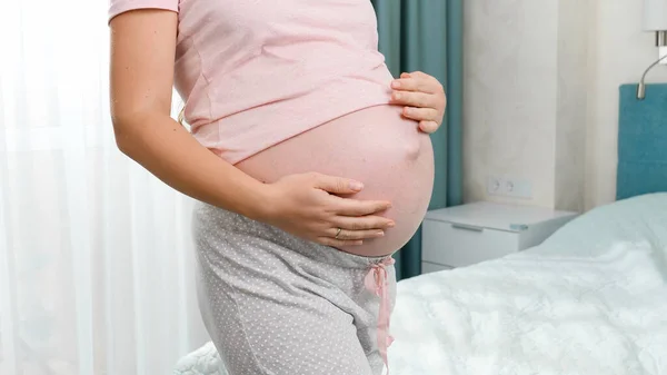 Fechar-se de mulher grávida no pijama que está na grande janela e acariciando a grande barriga. Conceito de felicidade durante a gravidez e esperando bebê — Fotografia de Stock