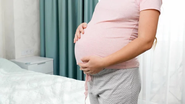 Närbild av gravida kvinna stroking och hålla stor mage vid fönstret i sovrummet. Begreppet lycka under graviditeten och väntar barn — Stockfoto