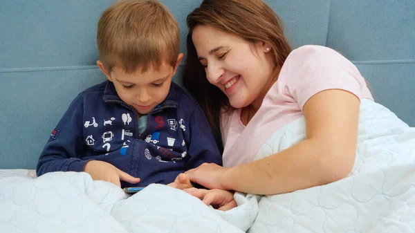 Retrato de feliz madre sonriente con pequeño hijo pequeño jugando en la tableta mientras está acostado en la cama —  Fotos de Stock