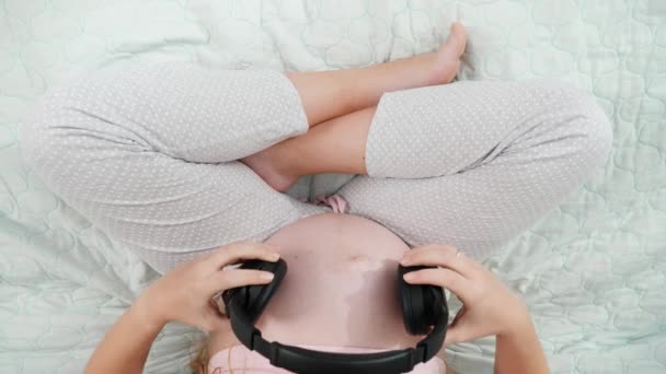 Κοντινό πλάνο της εγκύου γυναίκας με μεγάλη κοιλιά κάθεται στο κρεβάτι και βάζοντας μεγάλα ακουστικά στην κοιλιά της. Έννοια της πρώιμης μουσικής, της τέχνης και της δημιουργικότητας — Αρχείο Βίντεο