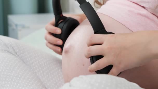 CLoseup jonge zwangere vrouw zet koptelefoon met klassieke muziek op haar grote buik. Begrip vroege muziek, kunst en creativiteit — Stockvideo