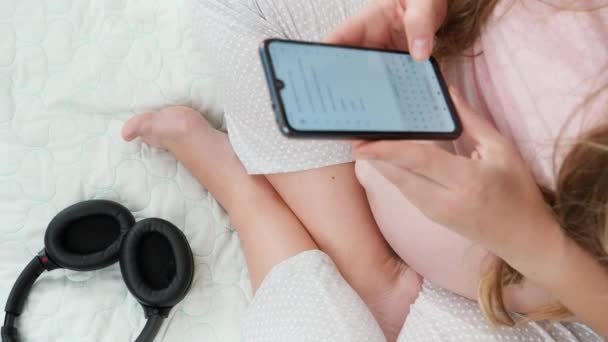 Gros plan de femmes enceintes assis sur le lit et tapant un message sur smartphone après avoir écouté de la musique dans un casque. Concept de musique ancienne, art et développement de la créativité — Video