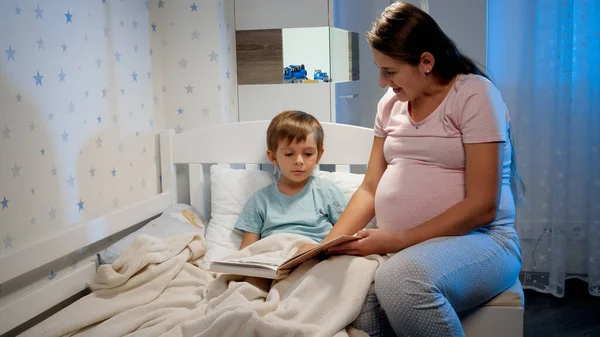 Hermosa madre embarazada readng libro de cuentos a su hijo mayor acostado en la cama por la noche antes de ir a dormir. Padres leyendo a los niños —  Fotos de Stock