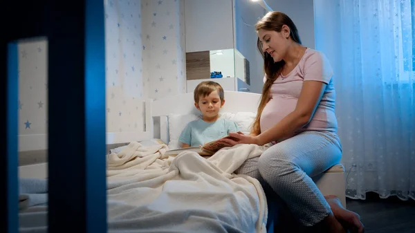 Carino bambino ragazzo ascoltando libro di fiabe sua madre incinta sta leggendo prima di andare a dormire la notte. Genitorialità e educazione dei figli — Foto Stock