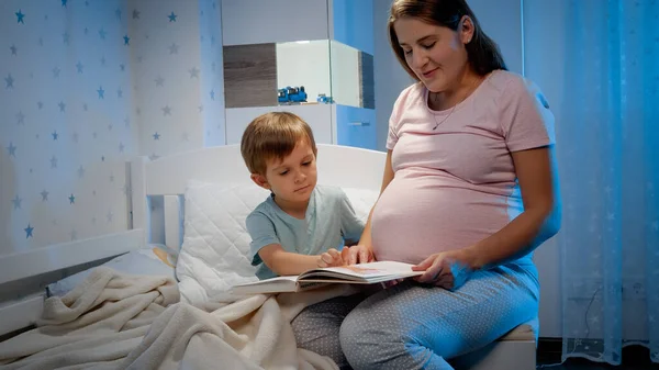 Niño sonriente con madre embarazada leyendo libro en la cama por la noche. Padres leyendo a sus hijos y pasando tiempo juntos —  Fotos de Stock