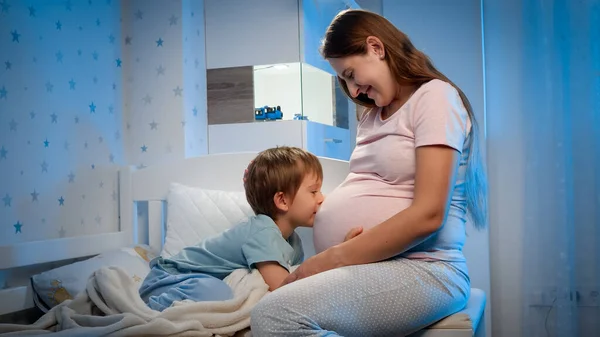 小さな男の子抱擁と話をします小さな生まれていない赤ん坊で大きな腹の中で妊娠中の母親が夜彼のベッドの横に座っている場合 — ストック写真
