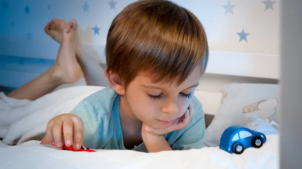 Portrait of little boy playing with two toy cars at bedtime. Toddler boy lying in bed and playing with cars — Stock Photo, Image