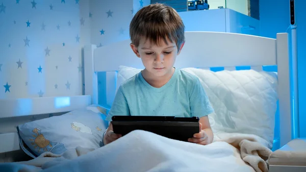 Portrait of little toddler boy sitting in bed at night and watching video on tablet computer. Little boy with gadget at night — Stock Photo, Image