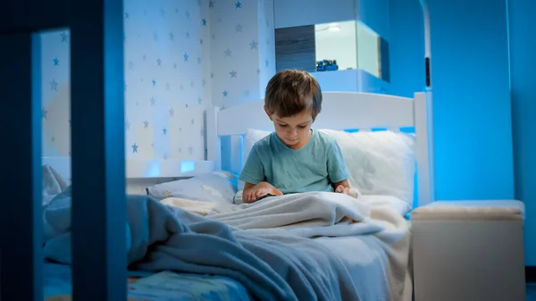 Little 5 years old boy using and playing on tablet computer before going to sleep at night. Child with gadget at evening — Stock Photo, Image