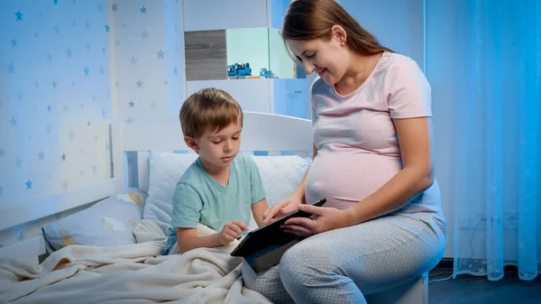 Jovem mãe grávida sorridente sentada ao lado de seu filho deitada na cama e assistindo vídeo no computador tablet — Fotografia de Stock