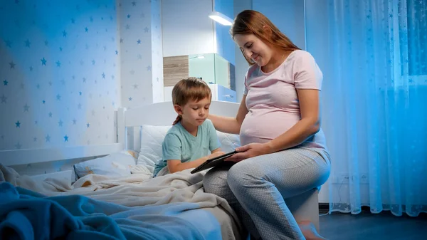 Маленький мальчик с молодой беременной матерью играет в игры и смотрит мультики на планшетном компьютере в постели перед сном — стоковое фото