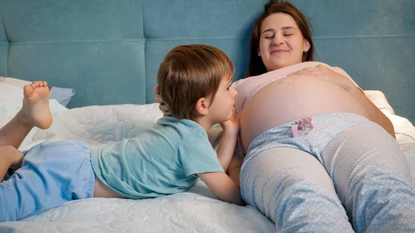 Retrato del niño pequeño en pijama acostado con la madre embarazada en la cama y hablando con el bebé nonato en el vientre grande. Niños cariñosos y cariñosos — Foto de Stock