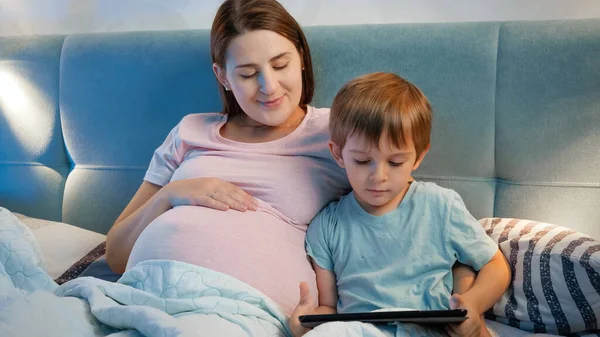 Portret van een zwangere jonge moeder die haar zoontje knuffelt, liggend in bed en spelend op een tablet computer voordat ze gaat slapen. — Stockfoto