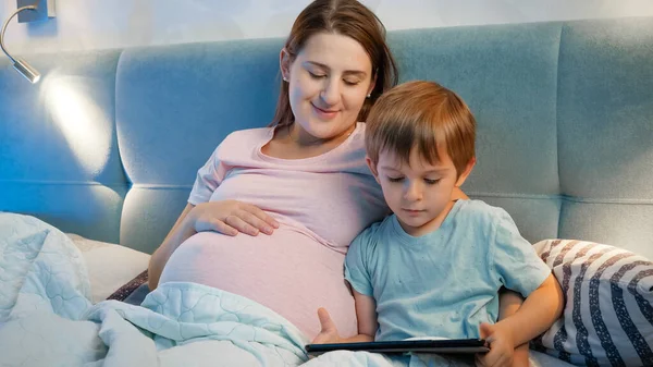 Menino criança usando tablet computador deitado com a mãe grávida na cama à noite — Fotografia de Stock