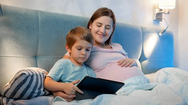 Семья лежит в постели и смотрит мультики на планшетном компьютере ночью. Маленький мальчик и беременная мать с помощью цифрового планшета в постели ночью — стоковое фото