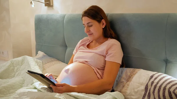 Mosolygó terhes nő pizsamában dolgozik tabletta és böngészés az interneten, mielőtt aludni az ágyban éjszaka — Stock Fotó