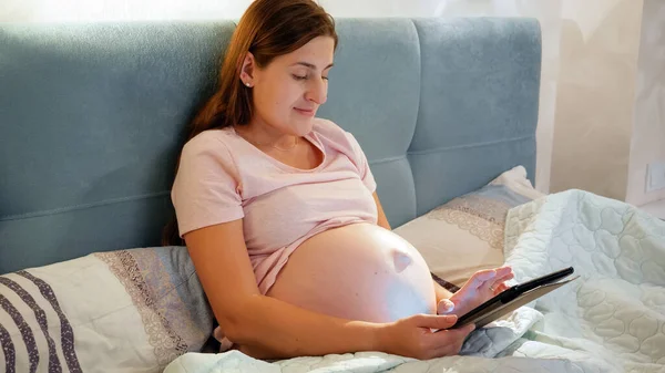 Mulher grávida sorrindo de pijama usando computador tablet e digitação mensagem enquanto deitado na cama à noite — Fotografia de Stock