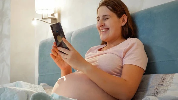 임신 한 여성이 잠옷을 입고 침대에 누운 채 미소짓고 스마트폰에서 비디오 컨퍼런스를 갖는 모습. 임신 한 젊은 여자가 비디오 전화로 이야기하는 모습 — 스톡 사진