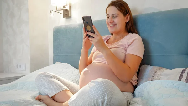 Mladá těhotná žena mluví na videohovoru, zatímco sedí v noci v posteli. Těhotná dívka mluví s přítelem nebo lékařem prostřednictvím videokonference na smartphonu. — Stock fotografie