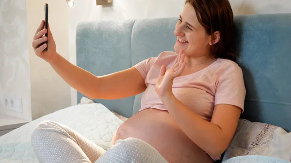 Felice donna incinta sorridente sdraiata a letto e parlare con un amico in videoconferenza. Ragazza incinta con videochiamata — Foto Stock