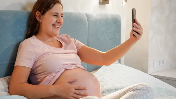 Bebek bekleyen gülümseyen hamile bir kadın yatakta oturup video konferansında doktor ya da arkadaşını arıyor. Akıllı telefon kullanan hamile bir kadın. — Stok fotoğraf