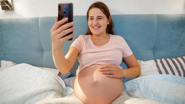 Lykkelig smilende, gravid kvinne som lager selfie på smarttelefon før hun legger seg om kvelden – stockfoto