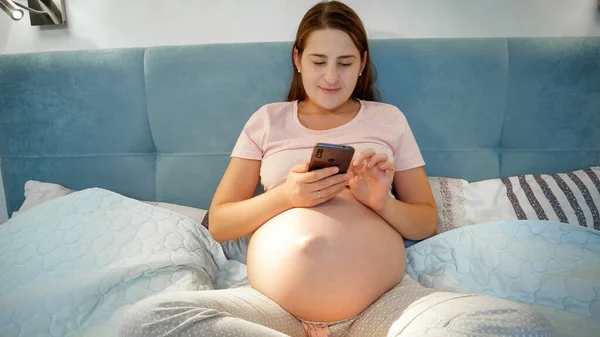 Pijamalı hamile bir kadın yatakta uzanıyor ve akıllı telefon kullanıyor. Hamile kadın ve telefon geceleri yatakta uzanıyor. — Stok fotoğraf