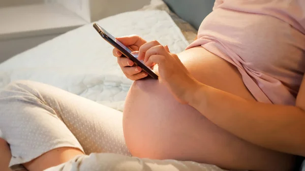 Close-up de mulher grávida com grande mensagem de digitação da barriga e navegação na internet no smartphone na cama à noite — Fotografia de Stock