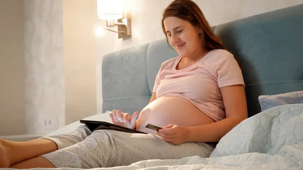 Leende gravid kvinna gör online shopping på surfplatta dator innan du går och lägger dig på natten. — Stockfoto