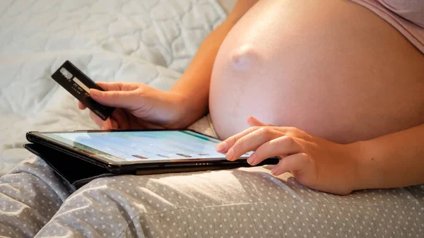 Close-up de mulher grávida com grande bely segurando cartão de crédito e fazer compras on-line em e-store no computador tablet à noite — Fotografia de Stock