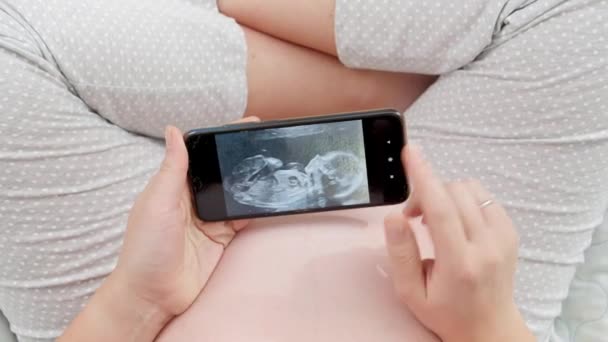 침대에 앉아 스마트폰으로 태어나지 않은 아기의 초음파 영상을 보고 있는 임신부의 모습 — 비디오