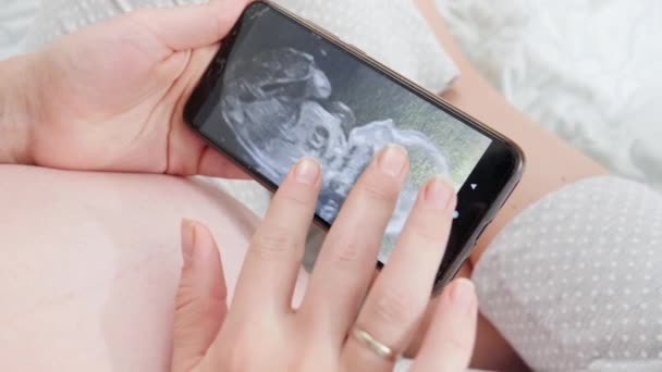 Nærbillede af gravid kvinde, der holder smartphone og ser på ultralydsbilledet af hendes ufødte baby i maven. Begrebet forventer baby, graviditet og sundhedspleje. – Stock-video