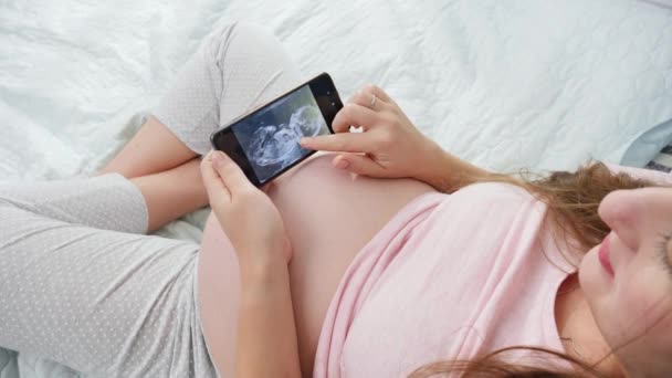 Jonge zwangere vrouw in pyjama bladert door ultrasoon beeld van haar ongeboren baby. Concept van het verwachten van baby, zwangerschap en gezondheidszorg. — Stockvideo