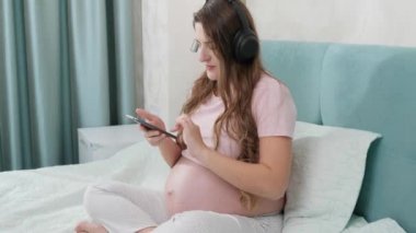 Mutlu gülümseyen hamile kadın kulaklıkla müzik dinliyor ve evde yatarken akıllı telefon kullanıyor.