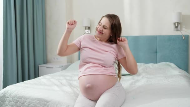 Щаслива весела вагітна жінка танцює і слухає музику, сидячи в ліжку вранці. Концепція щастя під час вагітності та очікування дитини — стокове відео