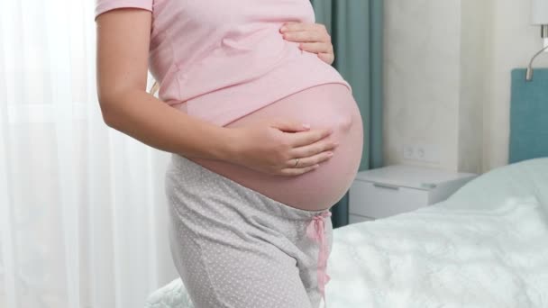 Fechar-se de mulher grávida no pijama que está na grande janela e acariciando a grande barriga. Conceito de felicidade durante a gravidez e esperando bebê — Vídeo de Stock