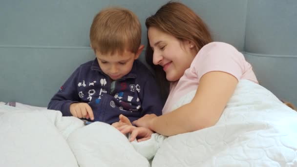 Retrato de mãe sorridente feliz com o pequeno filho criança brincando no computador tablet enquanto deitado na cama — Vídeo de Stock