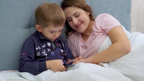 Menino criança com a jovem mãe deitada na cama de manhã e jogando jogos no computador tablet. — Vídeo de Stock