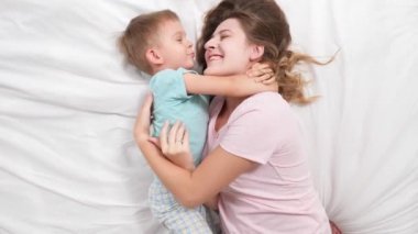 Pijamalı, gülümseyen annesini kucaklayan beyaz çarşaflı küçük bir çocuğun portresi. Ebeveynlik kavramı, sevgi dolu çocuklar ve aile mutluluğu.
