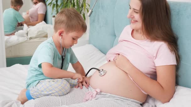 Doktorda oynayan sevimli çocuk ve steteskopla hamile annenin kalp atışlarını dinliyor. Hamilelik süresince sağlık ve tıbbi muayene kavramı — Stok video