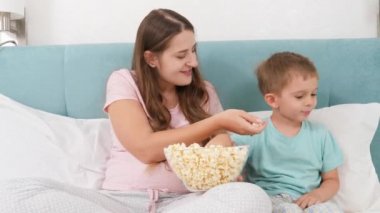 Küçük bir çocuk ve pijamalı genç bir anne hafta sonu yatağa uzanıp büyük kaseden patlamış mısır yiyor. Neşeli çocuklar ve aile mutluluğu kavramı.
