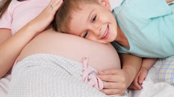 Милий маленький хлопчик у піжамі слухає ненароджену дитину у вагітному материнському животі, що лежить на ліжку. Концепція люблячих дітей і сімейного щастя очікує дитину — стокове відео