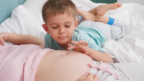 Feliz niño sonriente en pijama acariciando el vientre grande de la madre embarazada y hablando con ella. Concepto de amar a los niños y la felicidad familiar esperando al bebé — Vídeos de Stock