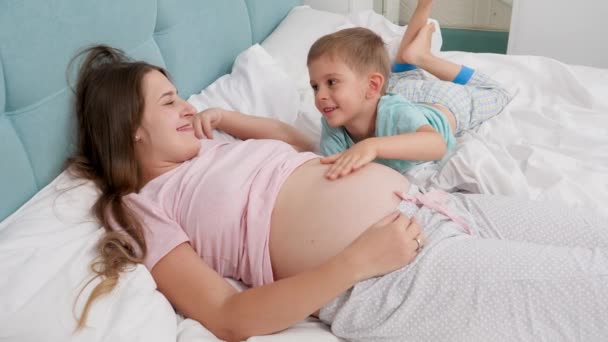 Feliz sorrindo mulher grávida com menino em pijama lyin na cama e falando. Conceito de crianças amorosas e felicidade familiar esperando bebê — Vídeo de Stock