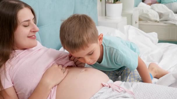 Lindo chico adorable besando y escuchando el vientre grande de su madre embarazada acostada en la cama. Concepto de amar a los niños y la felicidad familiar esperando al bebé — Vídeos de Stock