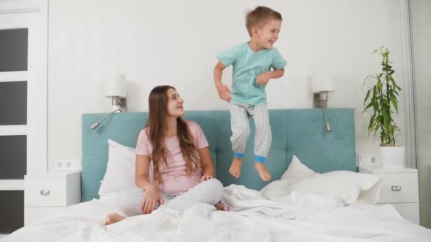 Bella giovane donna in pigiama guardando il suo piccolo figlio bambino che salta sul suo letto al mattino. Concetto di bambini allegri e felicità familiare. — Video Stock